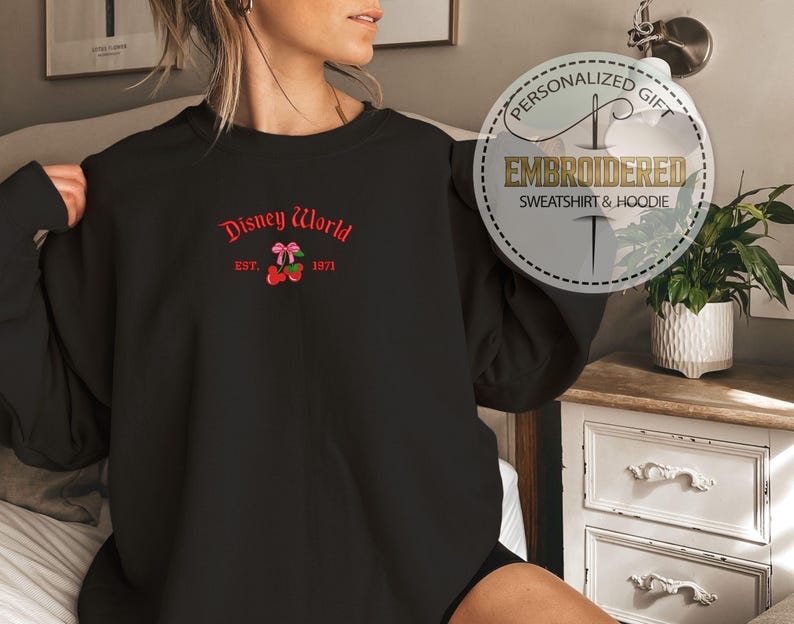May include: Black sweatshirt with embroidered "Disney World" text in red, along with a cherry and bow design. The text "EST. 1971" is also included. The sweatshirt is a personalized gift.