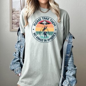 Op de afbeelding: Lichtgroen t-shirt met een cirkelvormige afbeelding met een ruimteschip, twee ontvoerde figuren en de tekst "PLEASE TAKE THEM" en "NO KINGS IN AMERICA". Het ontwerp gebruikt tinten rood, oranje, geel en blauw.
