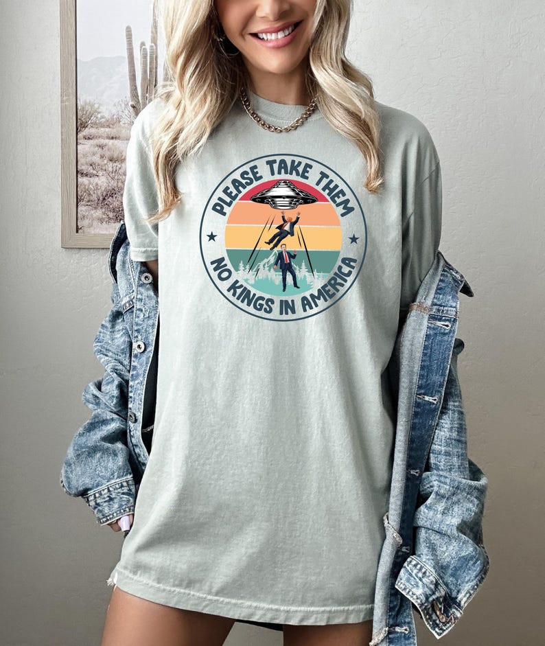 Op de afbeelding: Lichtgroen t-shirt met een cirkelvormige afbeelding met een ruimteschip, twee ontvoerde figuren en de tekst "PLEASE TAKE THEM" en "NO KINGS IN AMERICA". Het ontwerp gebruikt tinten rood, oranje, geel en blauw.