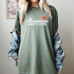 May include: Olive green t-shirt with a white graphic of a sailboat, a setting sun, and the text "AMITY 1975." The shirt has a relaxed fit and is made of a soft material. The shirt is a casual wear item.