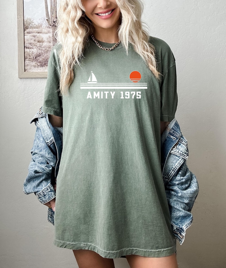 May include: Olive green t-shirt with a white graphic of a sailboat, a setting sun, and the text "AMITY 1975." The shirt has a relaxed fit and is made of a soft material. The shirt is a casual wear item.