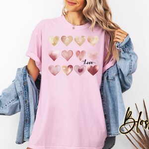 Puede incluir: Una camiseta rosa con un dise&ntilde;o de acuarela de corazones en tonos rosa y dorado. Los corazones est&aacute;n dispuestos en una cuadr&iacute;cula y la palabra "Love" est&aacute; escrita en letra negra debajo de los corazones.