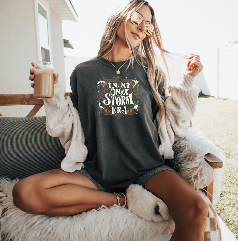 May include: A woman wearing a dark gray t-shirt with the text "In My Onyx Storm Era" in a vintage font. The shirt has a distressed look and features a small star and a few stylized dragons.
