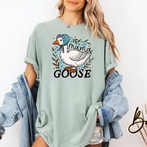 Puede incluir: Una camiseta azul claro con un gr&aacute;fico de un ganso con un gorro azul y rodeado de flores. El texto "Mama Goose" est&aacute; impreso en la camiseta.