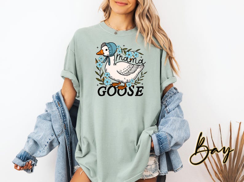 Puede incluir: Una camiseta azul claro con un gr&aacute;fico de un ganso con un gorro azul y rodeado de flores. El texto "Mama Goose" est&aacute; impreso en la camiseta.