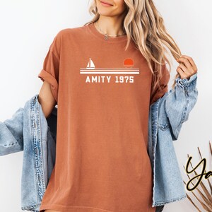 May include: A rust-colored t-shirt with the text "AMITY 1975" in white, above a graphic of a sailboat and a setting sun. The shirt is worn with a light blue denim jacket.