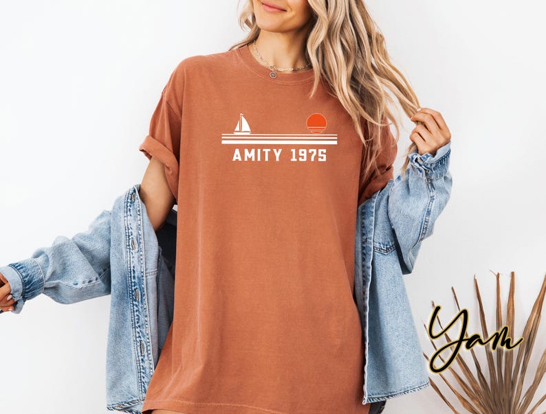 May include: A rust-colored t-shirt with the text "AMITY 1975" in white, above a graphic of a sailboat and a setting sun. The shirt is worn with a light blue denim jacket.