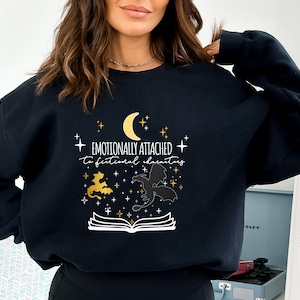 May include: Black sweatshirt with a white graphic design that says "Emotionally attached to fictional characters". The graphic also includes a crescent moon, stars, and two dragons.