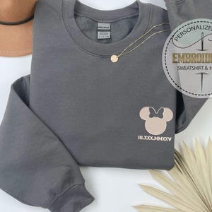 May include: A grey sweatshirt featuring a light brown Minnie Mouse design and Roman numerals. A gold necklace and a brown hat are also visible. The sweatshirt has the text "PERSONALIZED GIFT EMBROIDERED SWEATSHIRT & HOODIE" on a circular sticker.