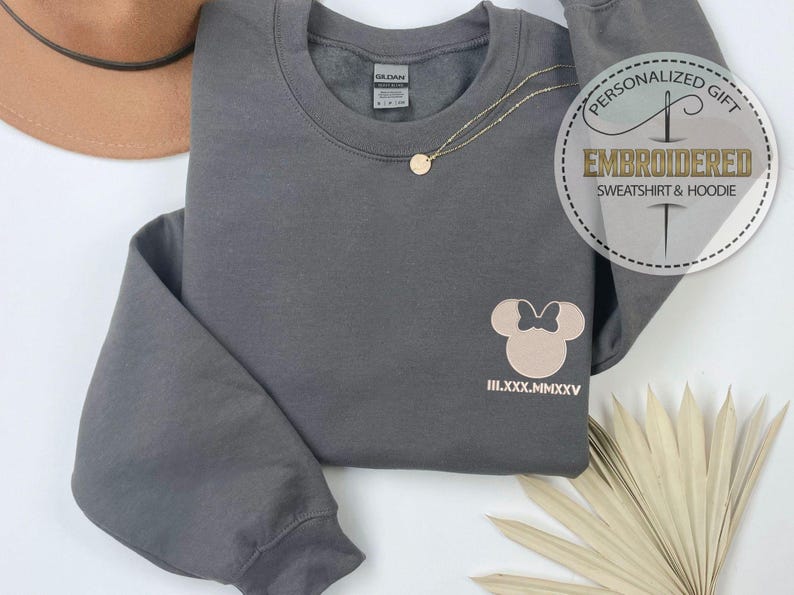 May include: A grey sweatshirt featuring a light brown Minnie Mouse design and Roman numerals. A gold necklace and a brown hat are also visible. The sweatshirt has the text "PERSONALIZED GIFT EMBROIDERED SWEATSHIRT & HOODIE" on a circular sticker.