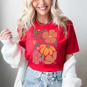 May include: Red t-shirt with a floral design featuring orange, brown, and pink flowers. The flowers are stylized and have a retro feel.