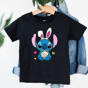 May include: Black t-shirt with a blue Stitch character wearing bunny ears and holding a colorful Easter egg. The character is surrounded by pink and yellow hearts.