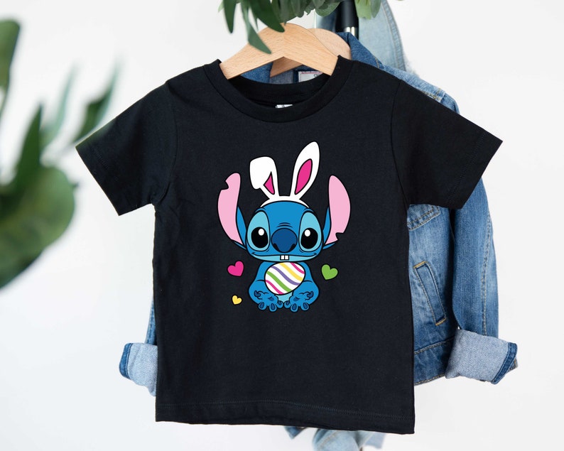 May include: Black t-shirt with a blue Stitch character wearing bunny ears and holding a colorful Easter egg. The character is surrounded by pink and yellow hearts.