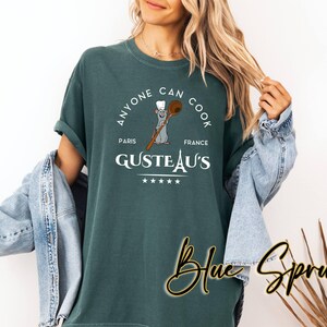 May include: Teal t-shirt with a circular design. The design includes the text "ANYONE CAN COOK", "GUSTEAU'S PARIS FRANCE", and a cartoon rat holding a wooden spoon. The words "Blue Spruce" are written in cursive.