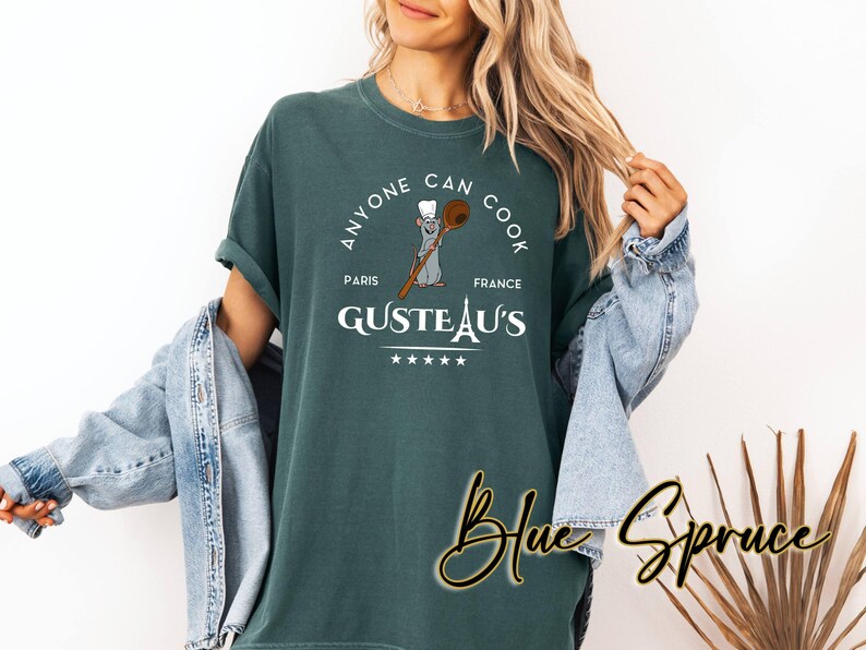 May include: Teal t-shirt with a circular design. The design includes the text "ANYONE CAN COOK", "GUSTEAU'S PARIS FRANCE", and a cartoon rat holding a wooden spoon. The words "Blue Spruce" are written in cursive.