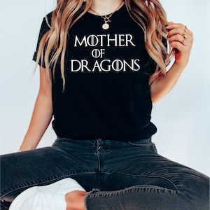 Op de afbeelding: Een zwart T-shirt met witte tekst die "Mother of Dragons" zegt. Het shirt wordt gedragen door een persoon die op een rode stoel zit.