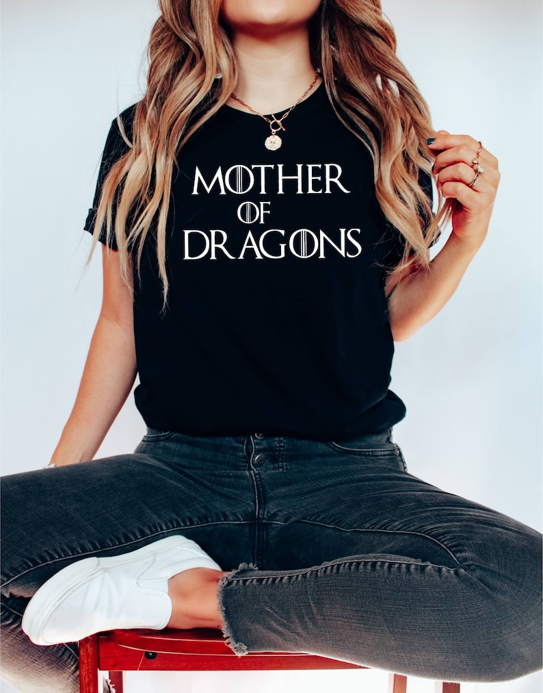 Op de afbeelding: Een zwart T-shirt met witte tekst die "Mother of Dragons" zegt. Het shirt wordt gedragen door een persoon die op een rode stoel zit.