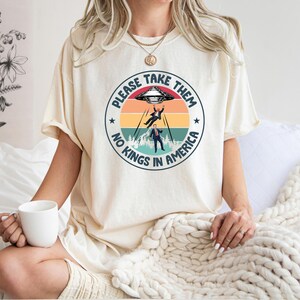 Op de afbeelding: Cr&egrave;mekleurig T-shirt met een cirkelvormige afbeelding van een buitenaardse ontvoering. De tekst luidt "PLEASE TAKE THEM" en "NO KINGS IN AMERICA". Het ontwerp bevat een retro zonsondergang en een UFO.