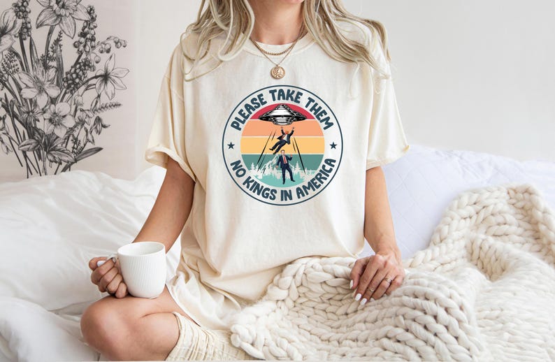 Op de afbeelding: Cr&egrave;mekleurig T-shirt met een cirkelvormige afbeelding van een buitenaardse ontvoering. De tekst luidt "PLEASE TAKE THEM" en "NO KINGS IN AMERICA". Het ontwerp bevat een retro zonsondergang en een UFO.