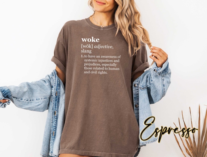 May include: A brown t-shirt with the word "woke" defined in white text. The definition reads: "[wōk] adjective, slang 1. to have an awareness of systemic injustices and prejudices, especially those related to human and civil rights."