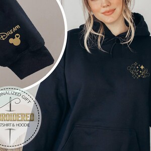 May include: Black hoodie with gold embroidered design. The design includes a Mickey Mouse silhouette and the word "Dream" on the sleeve. The hoodie has a front pocket and a drawstring hood. The image also includes a circular graphic that says "Personalized Gift" and "Embroidered Sweatshirt & Hoodie."