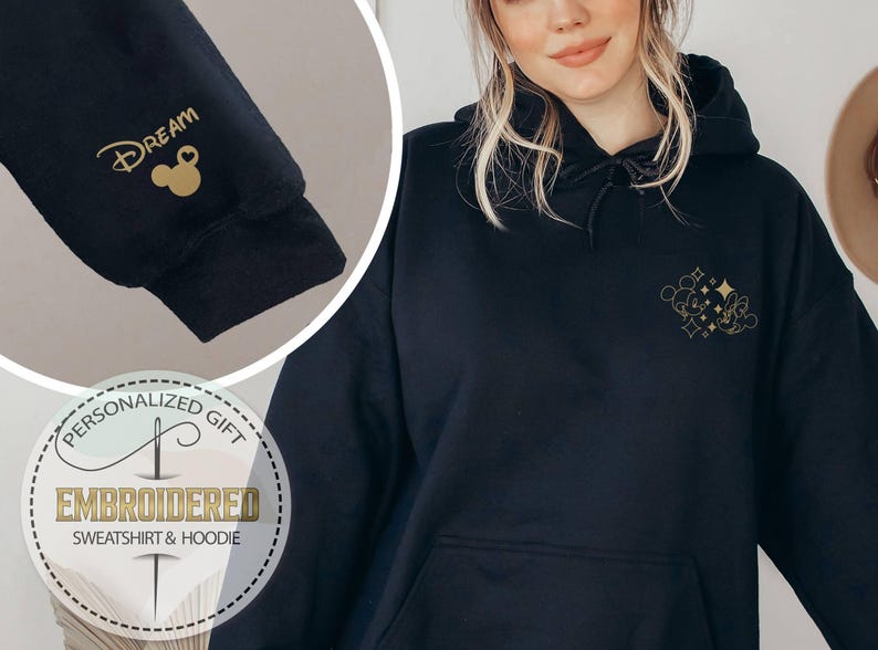 May include: Black hoodie with gold embroidered design. The design includes a Mickey Mouse silhouette and the word "Dream" on the sleeve. The hoodie has a front pocket and a drawstring hood. The image also includes a circular graphic that says "Personalized Gift" and "Embroidered Sweatshirt & Hoodie."