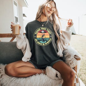 Op de afbeelding: Donkergrijze sweatshirt met een cirkelvormige afbeelding met een UFO, een ontvoerd persoon en de tekst "PLEASE TAKE THEM, NO KINGS IN AMERICA". Het ontwerp maakt gebruik van een kleurenschema van zonsondergang. De sweatshirt wordt door een persoon gedragen.