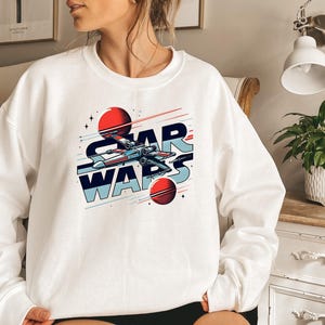 Puede incluir: Sudadera blanca con un gr&aacute;fico de una nave espacial volando por el espacio con planetas y el texto "STAR WARS" en azul y rojo.