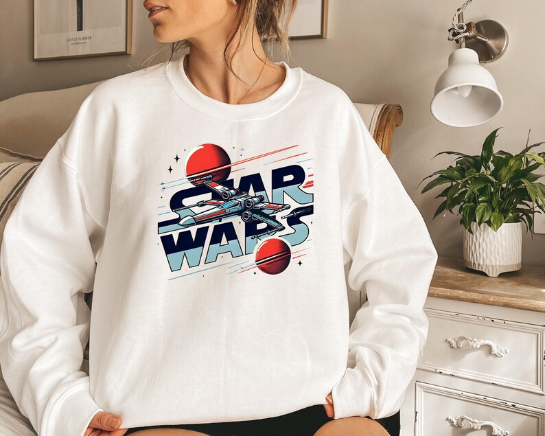 Puede incluir: Sudadera blanca con un gr&aacute;fico de una nave espacial volando por el espacio con planetas y el texto "STAR WARS" en azul y rojo.