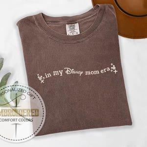 May include: A brown Comfort Colors t-shirt with the phrase "in my Disney mom era" in white script. The design includes Mickey Mouse head icons. A brown hat is visible in the background.