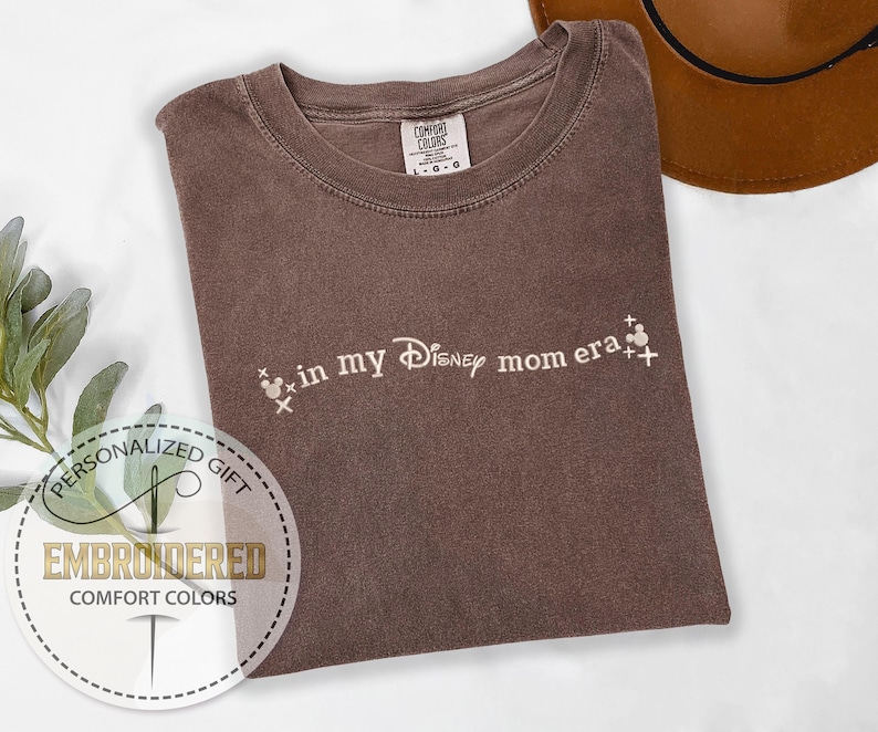 May include: A brown Comfort Colors t-shirt with the phrase "in my Disney mom era" in white script. The design includes Mickey Mouse head icons. A brown hat is visible in the background.