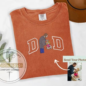 May include: Burnt orange Comfort Colors t-shirt with the word "DAD" embroidered in white block letters. The "A" is replaced with an illustration of a person helping a child. The shirt has a tag that says "COMFORT COLORS".