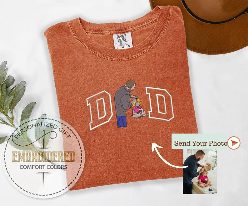 May include: Burnt orange Comfort Colors t-shirt with the word "DAD" embroidered in white block letters. The "A" is replaced with an illustration of a person helping a child. The shirt has a tag that says "COMFORT COLORS".