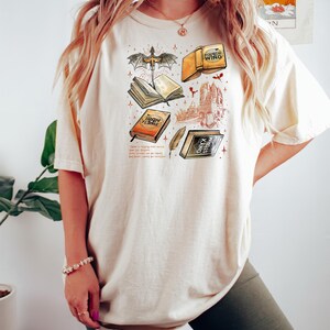 May include: A cream-coloured t-shirt with a fantasy-themed graphic. The design includes illustrations of books, a dragon, a feather, and a castle. The text on the shirt reads "Fourth Wing", "Iron Flame", and "Onyx Storm".