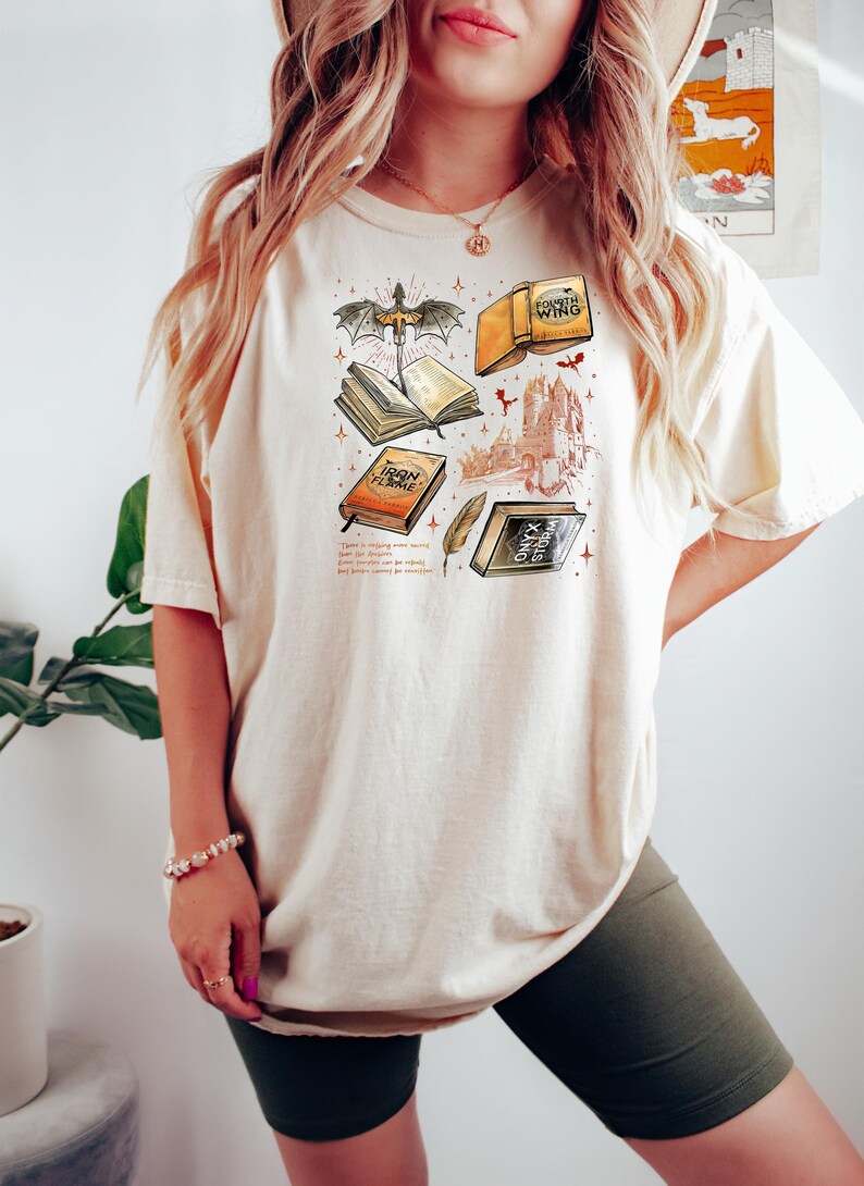 May include: A cream-coloured t-shirt with a fantasy-themed graphic. The design includes illustrations of books, a dragon, a feather, and a castle. The text on the shirt reads "Fourth Wing", "Iron Flame", and "Onyx Storm".