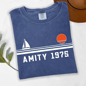 May include: A navy blue t-shirt with a white sailboat graphic, a white horizontal stripe, and the text "AMITY 1975". An orange sun graphic is above the text. A brown hat is in the background.
