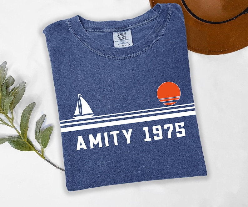May include: A navy blue t-shirt with a white sailboat graphic, a white horizontal stripe, and the text "AMITY 1975". An orange sun graphic is above the text. A brown hat is in the background.