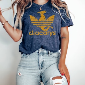 Pu&ograve; includere: Una t-shirt blu navy con una grafica di drago dorata invecchiata e il testo "dracarys" in oro. Il drago &egrave; in un design stilizzato a tre strisce.