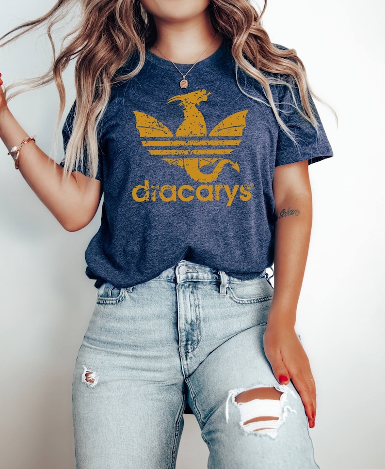 Pu&ograve; includere: Una t-shirt blu navy con una grafica di drago dorata invecchiata e il testo "dracarys" in oro. Il drago &egrave; in un design stilizzato a tre strisce.