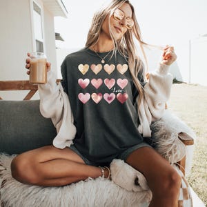 Puede incluir: Una camiseta gris antracita con un dise&ntilde;o de acuarela de corazones rosas y dorados. La camiseta tiene la palabra "Love" escrita en blanco en la parte delantera.