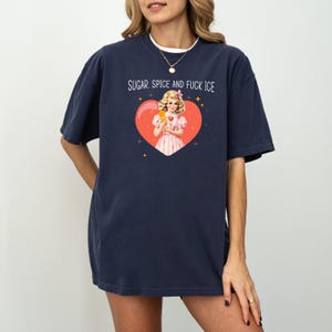 May include: Navy blue t-shirt featuring the text "SUGAR, SPICE AND FUCK ICE" above a heart-shaped graphic. The graphic shows a vintage illustration of a woman in a pink dress holding a popsicle. The shirt has a relaxed, oversized fit.