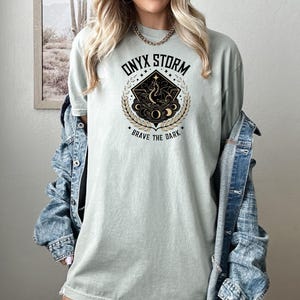 May include: A light green t-shirt with a black and gold graphic design featuring a dragon, stars, and moons. The graphic has the text "ONYX STORM" and "BRAVE THE DARK".