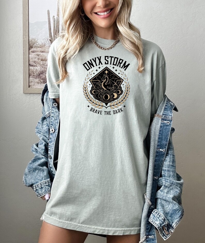 May include: A light green t-shirt with a black and gold graphic design featuring a dragon, stars, and moons. The graphic has the text "ONYX STORM" and "BRAVE THE DARK".