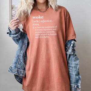 May include: A rust-colored t-shirt with the word "woke" and its definition printed in white. The definition is: "[wōk] adjective, slang 1. to have an awareness of systemic injustices and prejudices, especially those related to human and civil rights."