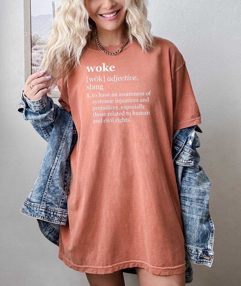 May include: A rust-colored t-shirt with the word "woke" and its definition printed in white. The definition is: "[wōk] adjective, slang 1. to have an awareness of systemic injustices and prejudices, especially those related to human and civil rights."
