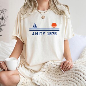 May include: Cream-colored t-shirt with a navy blue sailboat, orange sun, and the text "AMITY 1975." The shirt is a relaxed fit. The person is holding a white mug.