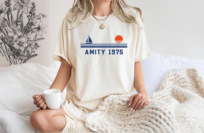 May include: Cream-colored t-shirt with a navy blue sailboat, orange sun, and the text "AMITY 1975." The shirt is a relaxed fit. The person is holding a white mug.
