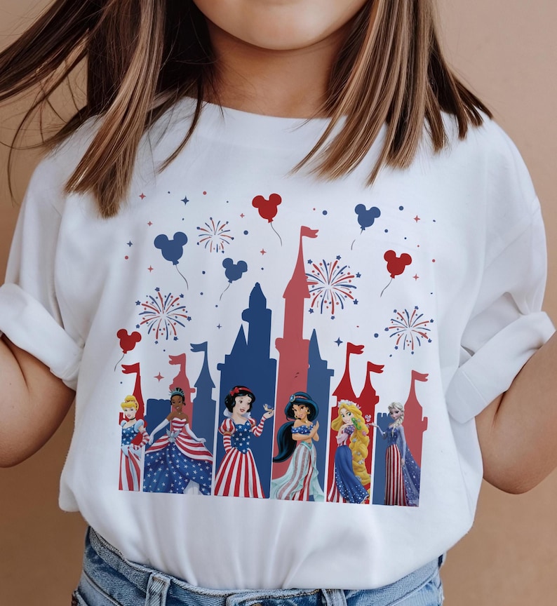 May include: White t-shirt featuring a patriotic castle design with red and blue accents, fireworks, and Mickey Mouse balloons. The shirt showcases various princess characters in festive attire, perfect for a themed celebration.