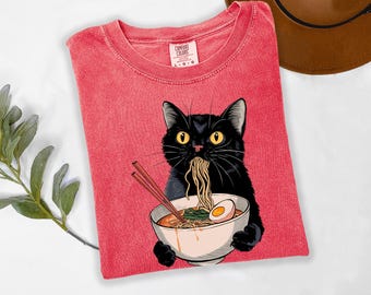 May include: Red Comfort Colors t-shirt featuring a black cat eating ramen from a white bowl. The cat has yellow eyes and is surrounded by noodles, chopsticks, and a boiled egg. The shirt is a size L-G.