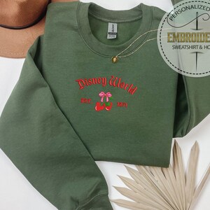 May include: An olive green sweatshirt with the embroidered text "Disney World" in red script, with a pink bow and cherries design. The sweatshirt also has the text "EST. 1971". A gold necklace and a brown hat are also visible.